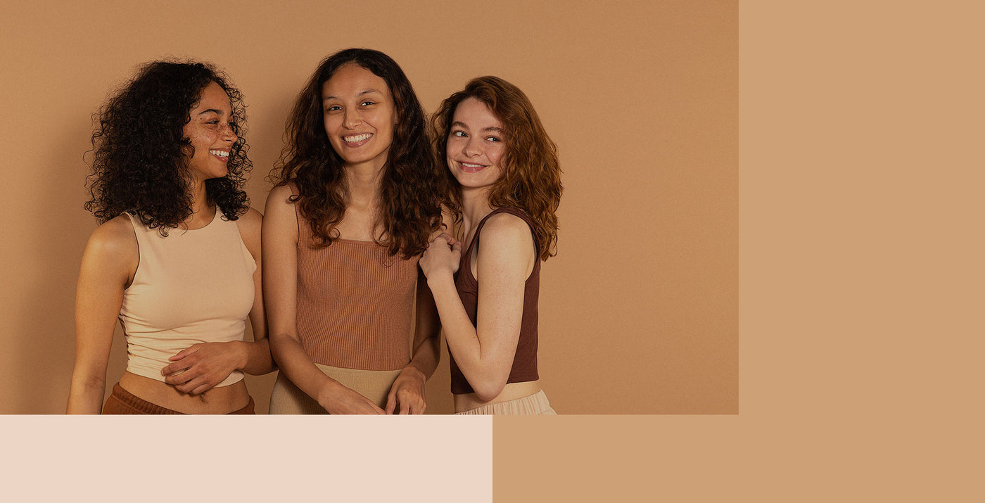 3 young women wearing neutral clothing against a neutral background smiling at eachother. 